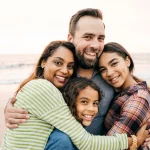 photo of man hugging his wife and two daughters as they all stand on a beach shoreline