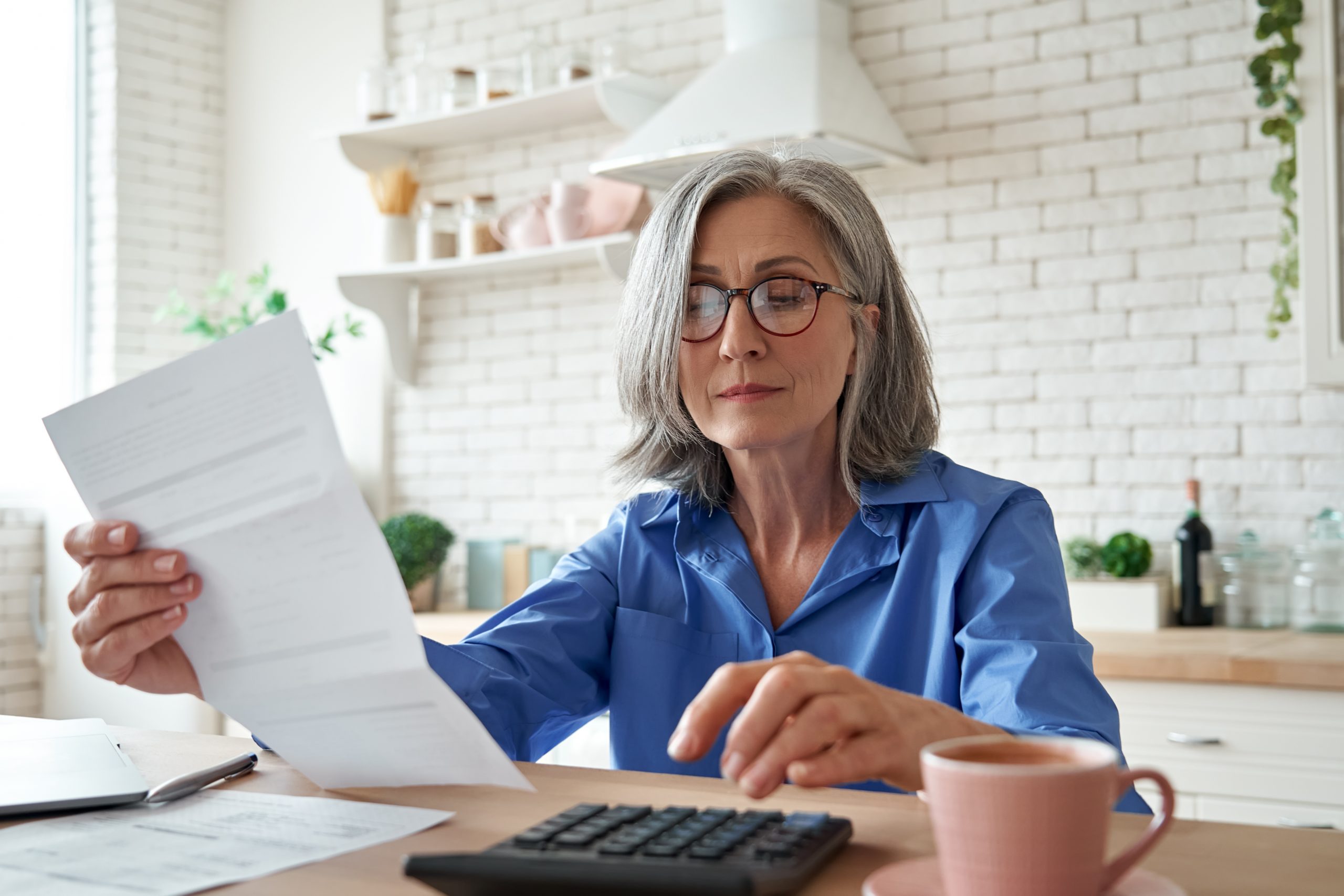 Senior mature business woman holding paper bill using calculator, old lady managing account finance, calculating money budget tax, planning banking loan debt pension payment sit at home kitchen table. Woman IRA planning, and planning for retirement with The Resource Center.